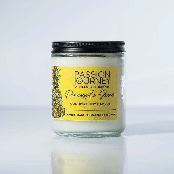 A clear glass candle with a black lid labeled 'Passion Journey - A Lifestyle Brand', with the scents listed as Amber, Sage, Pineapple. The candle is set against a white background. amber, sage, pineapple